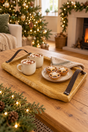 A rustic and made oak wooden serving tray with leather handles holds two white mugs of hot chocolate topped with marshmallows and cocoa powder, alongside a small plate of Christmas mince pies on a folded napkin, set on a wooden table in a cozy holiday living room.