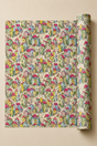 A roll of wrapping paper with a dense, colorful pattern of whimsical houses, trees, and paths on a cream background, shown partially unrolled against a solid beige backdrop.