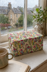 A rectangular gift box wrapped in Colourful Village Watercolour Wrapping Paper sits on a white wooden windowsill. Raindrops are visible on the window, with a mug, open book, and potted plant nearby.