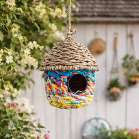 Colourful woven birdhouse hanging in a garden with flowers and trees.