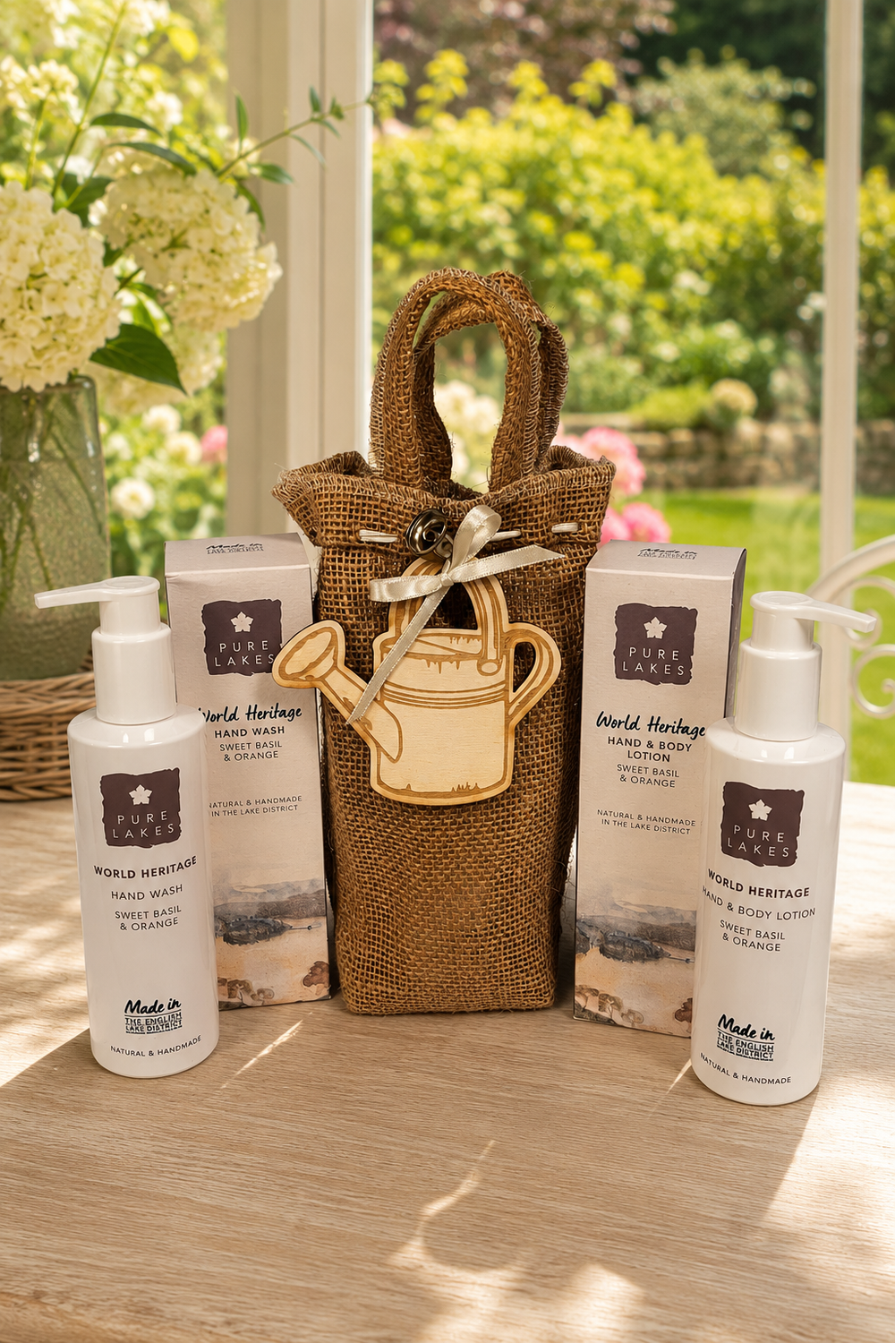 A gift set from World Heritage Hand & Body Care products arranged on a wooden table in front of a window with a garden view. The set includes a brown burlap tote bag with a wooden watering can charm, two white pump bottles, and two white boxes, all branded with the World Heritage logo.