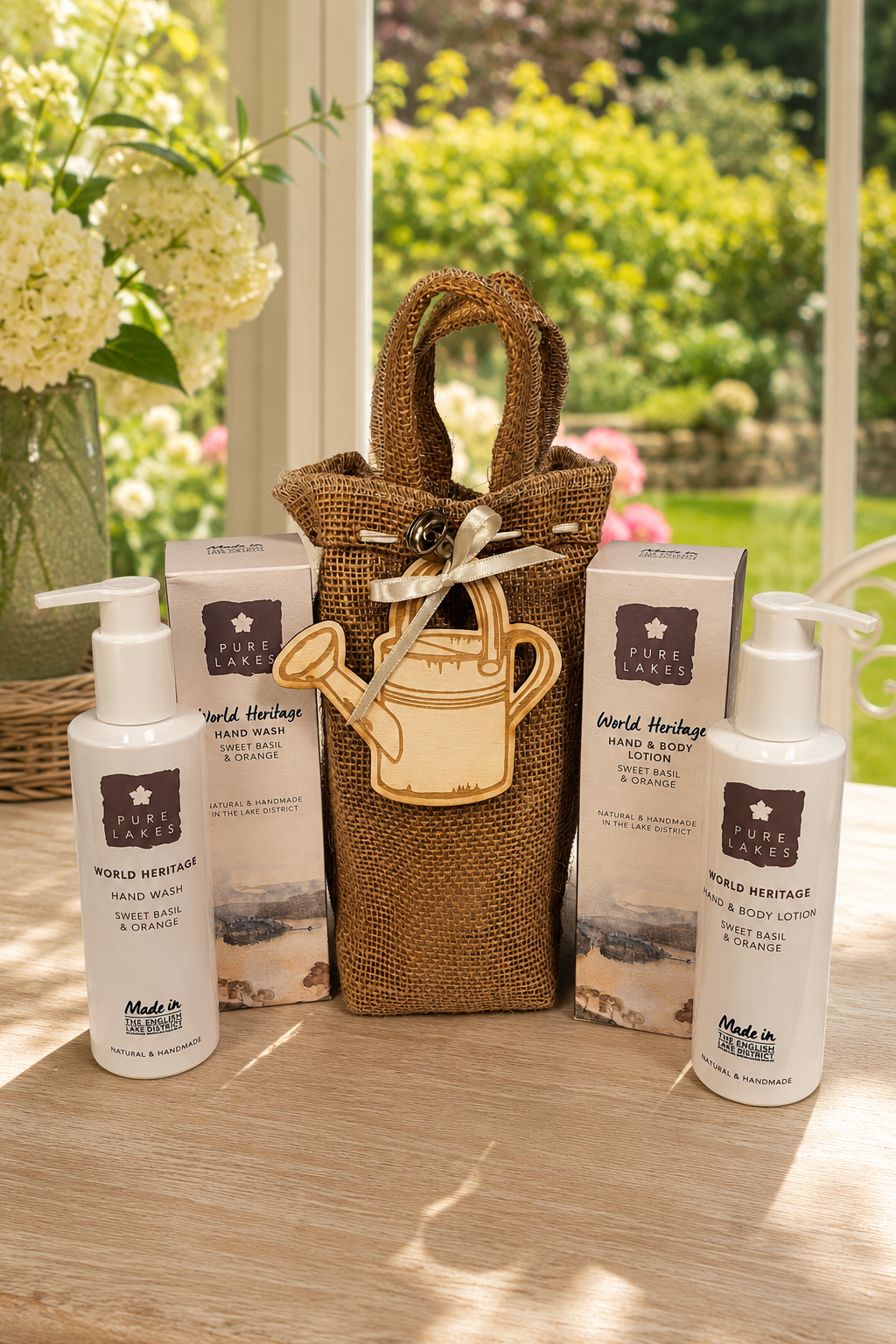 A gift set from World Heritage Hand & Body Care products arranged on a wooden table in front of a window with a garden view. The set includes a brown burlap tote bag with a wooden watering can charm, two white pump bottles, and two white boxes, all branded with the World Heritage logo.