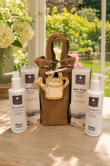 A gift set from World Heritage Hand & Body Care products arranged on a wooden table in front of a window with a garden view. The set includes a brown burlap tote bag with a wooden watering can charm, two white pump bottles, and two white boxes, all branded with the World Heritage logo.
