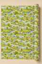 A roll of wallpaper featuring a repeating watercolor-style landscape pattern of green fields, trees, blue winding streams, and small white houses with orange roofs, shown against a warm beige background.