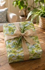 A gift box wrapped in a scenic landscape Water colour print wrapping paper tied with a burlap ribbon and rosemary sprig, set on a rustic wooden table with a cozy indoor background.