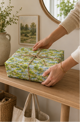 A person's hands wrapping a square gift box in a green and blue landscape paper, tied with a natural twine bow, set on a wooden table in a cozy room.