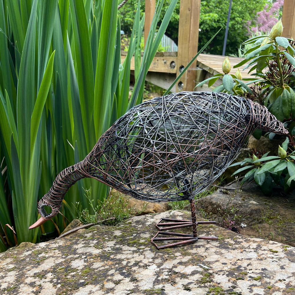 Decorative metal sculpture of a duck on a stone next to a pond with greenery