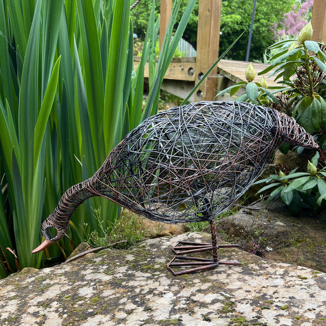 Decorative metal sculpture of a duck on a stone next to a pond with greenery