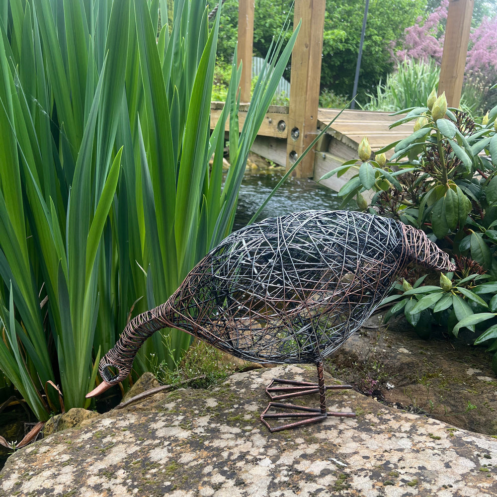 Decorative metal sculpture of a duck on a stone next to a pond with greenery