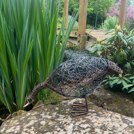 Decorative metal sculpture of a duck on a stone next to a pond with greenery