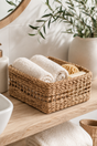 Woven basket with rolled towels on a wooden surface, with a plant and candle in the background.