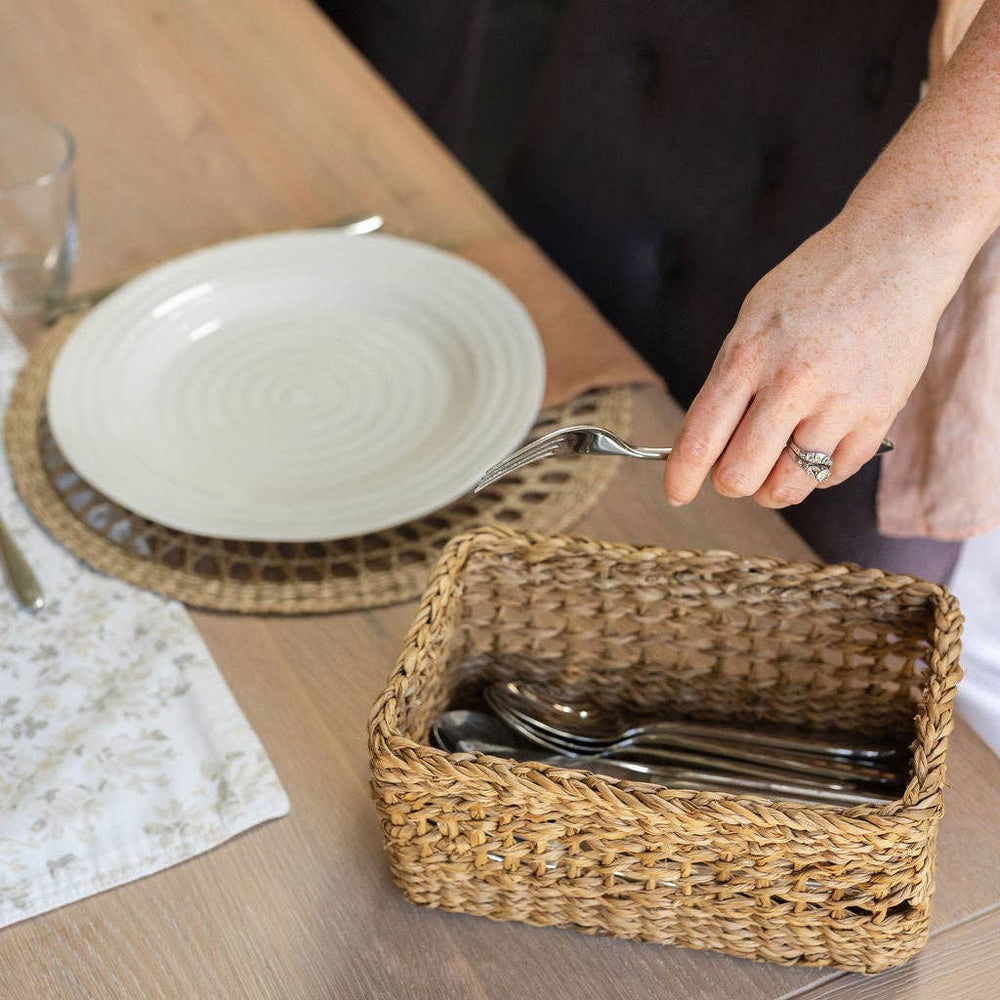 Person setting a table with a wicker basket containing cutlery, a white plate, and a glass.