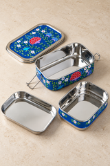 Rectangular stainless steel and enamel lunchbox, hand-painted in Kashmir with multicoloured floral decoration on a dark blue background. Includes two stacking containers, a shallow tray and a matching lid, providing three compartments for separating food. Metal side clips fasten the assembled lunchbox. Measures 18cm long × 12cm wide × 8cm high