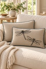 Two throw pillows with duck egg piped edges are styled on a cream sofa. The front pillow features a black printed dragonfly design, and a chunky cream knit throw blanket is draped over the sofa in a bright, sunlit living room setting.