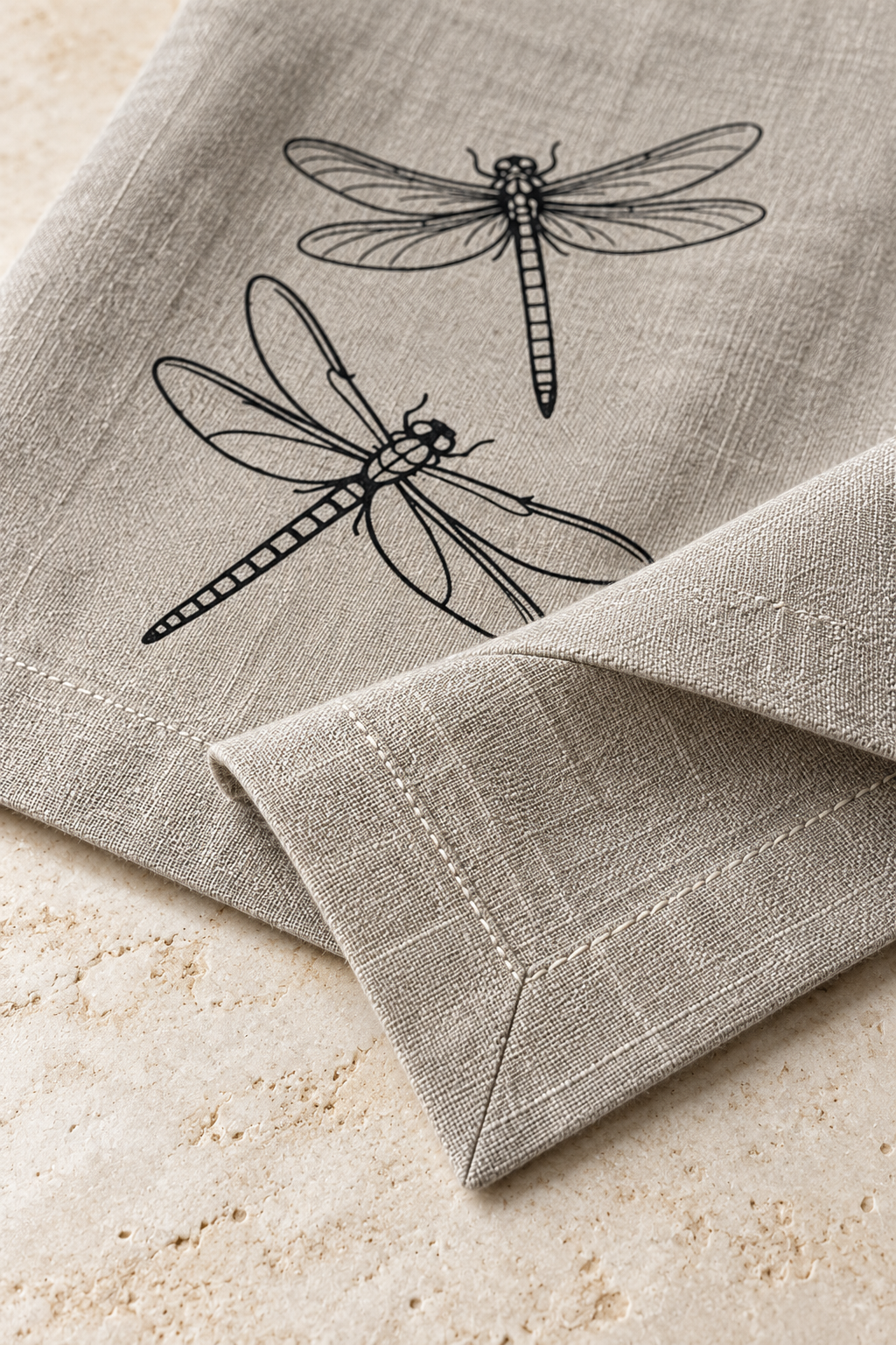 Close-up of a natural linen dragonfly napkin showing its mitred corner, double-fold hem and pale stitching.