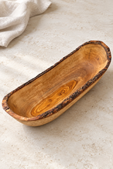 A rustic handcrafted wooden bowl with a natural bark edge, displayed on a light beige stone surface with a white linen cloth in the background.