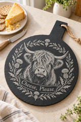 A round black slate cheese board with a handle, featuring a white engraved illustration of a Highland cow surrounded by thistles and the text 'Cheese, Please'. The board is styled on a beige tabletop with a knife, crackers, and a slice of cheese nearby.