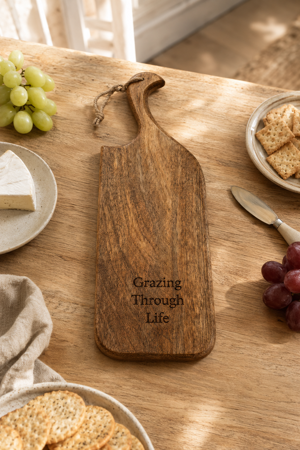 A wooden serving board with a handle and engraved text reading 'Grazing Through Life' on a wooden table, surrounded by food props like grapes, crackers, cheese, and a knife.