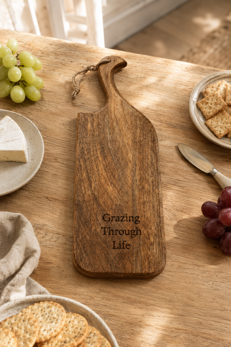 A wooden serving board with a handle and engraved text reading 'Grazing Through Life' on a wooden table, surrounded by food props like grapes, crackers, cheese, and a knife.