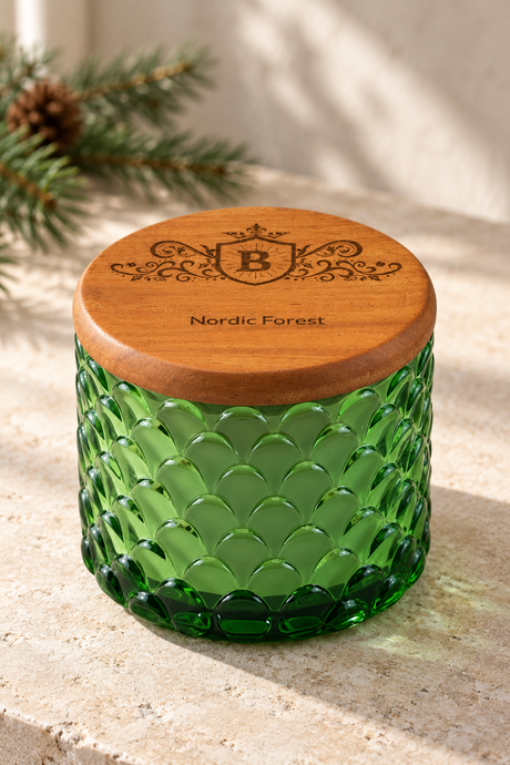 A green textured glass jar candle with a round wooden lid engraved with a crest and the words 'Nordic Forest', set on a stone surface with soft natural light and pine branches in the background.