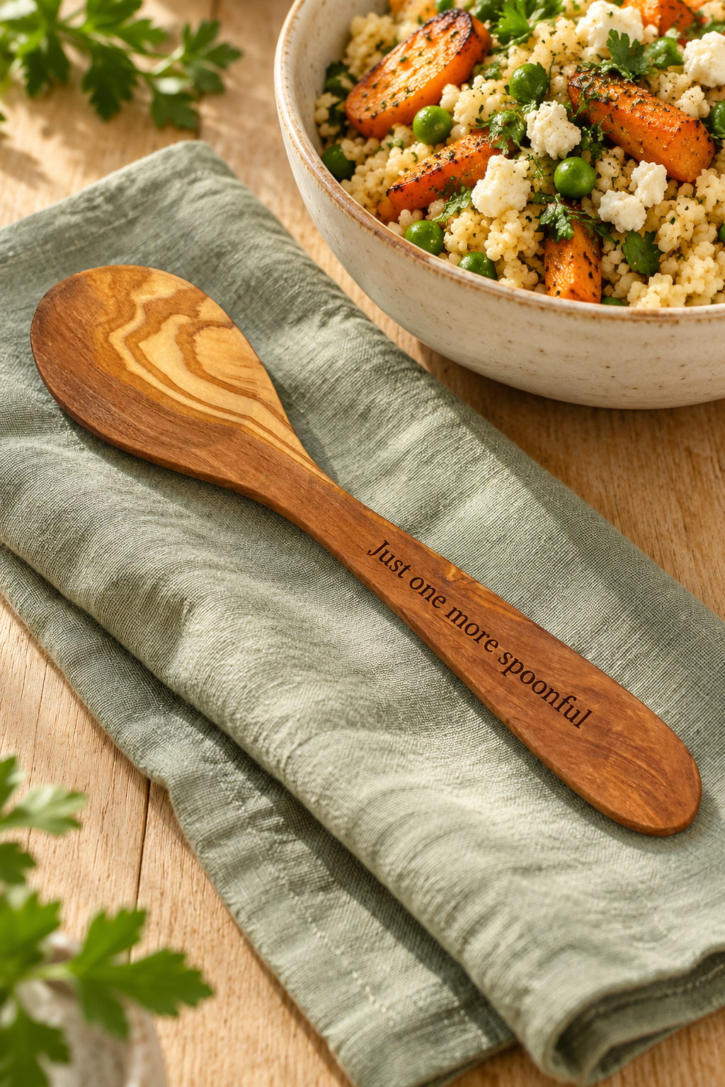 Olive wood serving spoon engraved with “Just one more spoonful”, resting on sage linen beside a bowl of couscous and roasted vegetables.