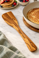 Olive wood spatula engraved “Flippin’ Good”, beside a frying pan, pancakes and fresh berries.