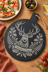 A round black slate serving board with a handle, featuring a detailed engraving of a stag surrounded by leaves and the words 'Woodland Feast'. The board is styled on a wooden table with a slice of pizza, olives, a glass of white wine with a lemon slice, and cherry tomatoes.