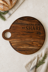 A round dark brown wooden serving board with a handle, featuring the engraved text 'ROUND HERE WE SHARE'. The board is styled in a flat-lay composition on a light beige textured surface, surrounded by a crusty loaf of bread, a beige cloth napkin, and rosemary sprigs.