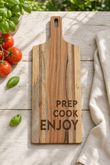 A rectangular wooden cutting board with a handle, featuring the engraved words 'PREP COOK ENJOY' in dark lettering. The board is styled on a light wooden table with fresh tomatoes, basil leaves, a green plant, and a beige cloth.
