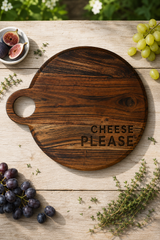 A round dark wood cheese board with a handle and the words 'CHEESE PLEASE' engraved on it, styled on a light wood tabletop with grapes, figs, herbs, and a small dish of sliced figs.