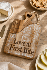A rustic wooden serving board with the engraved phrase 'Love at First Bite' and a heart-shaped handle, styled on a wooden table with cheese, crackers, and apple slices nearby.