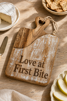 A rustic wooden serving board with the engraved phrase 'Love at First Bite' and a heart-shaped handle, styled on a wooden table with cheese, crackers, and apple slices nearby.