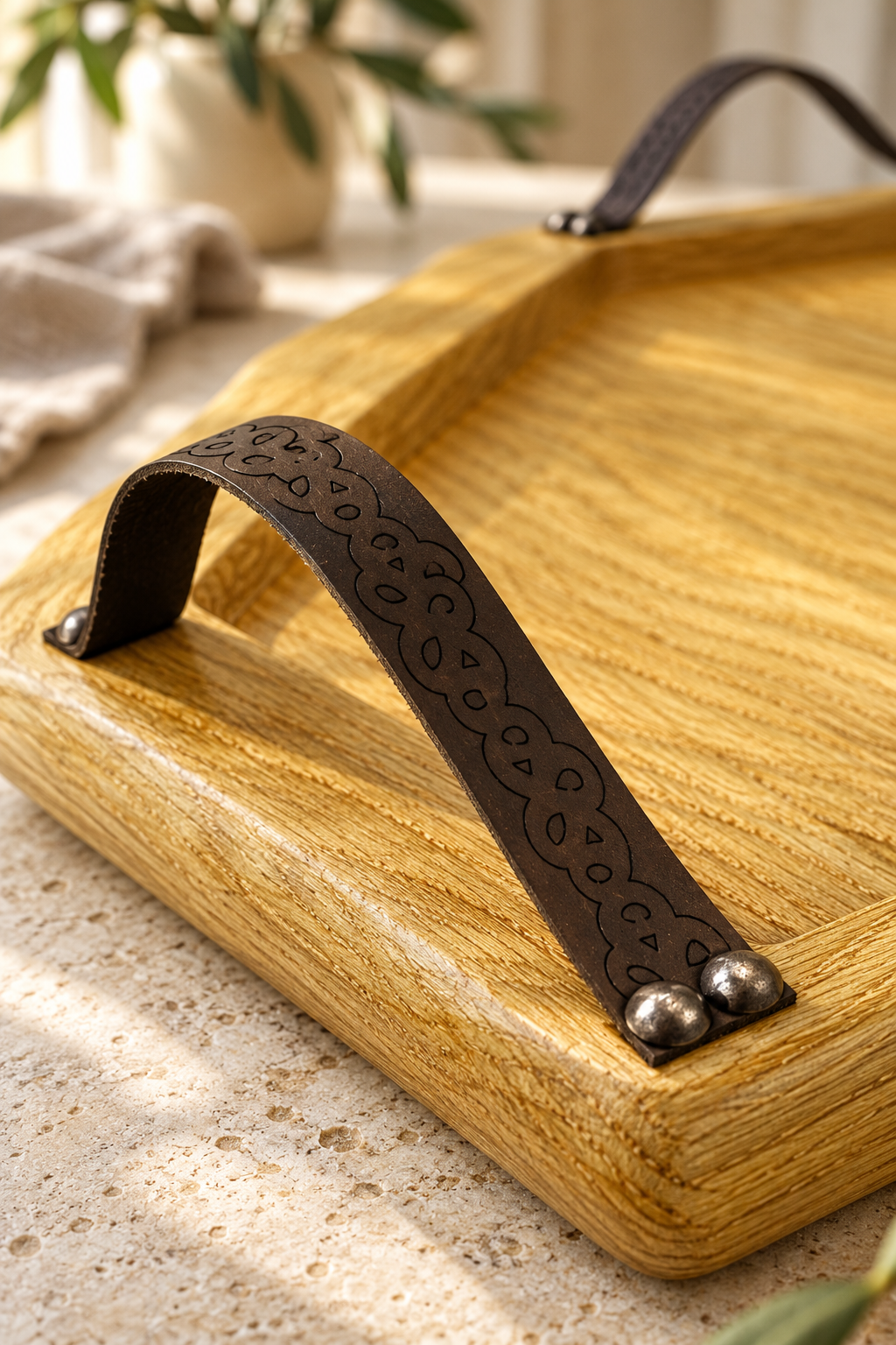 A close-up of a rectangular oak wood  serving tray with a dark brown leather handle featuring an embossed floral pattern and silver-tone metal rivets, set on a light stone surface.