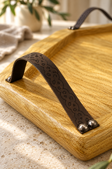 A close-up of a rectangular oak wood  serving tray with a dark brown leather handle featuring an embossed floral pattern and silver-tone metal rivets, set on a light stone surface.