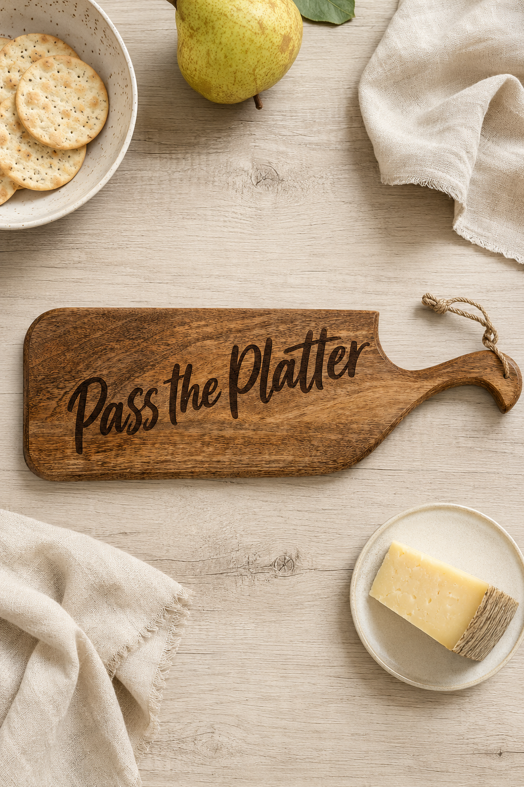 A wooden serving board engraved with the phrase 'Pass the Platter' in black script, styled on a light wood tabletop with a pear, crackers, cheese, and napkins.