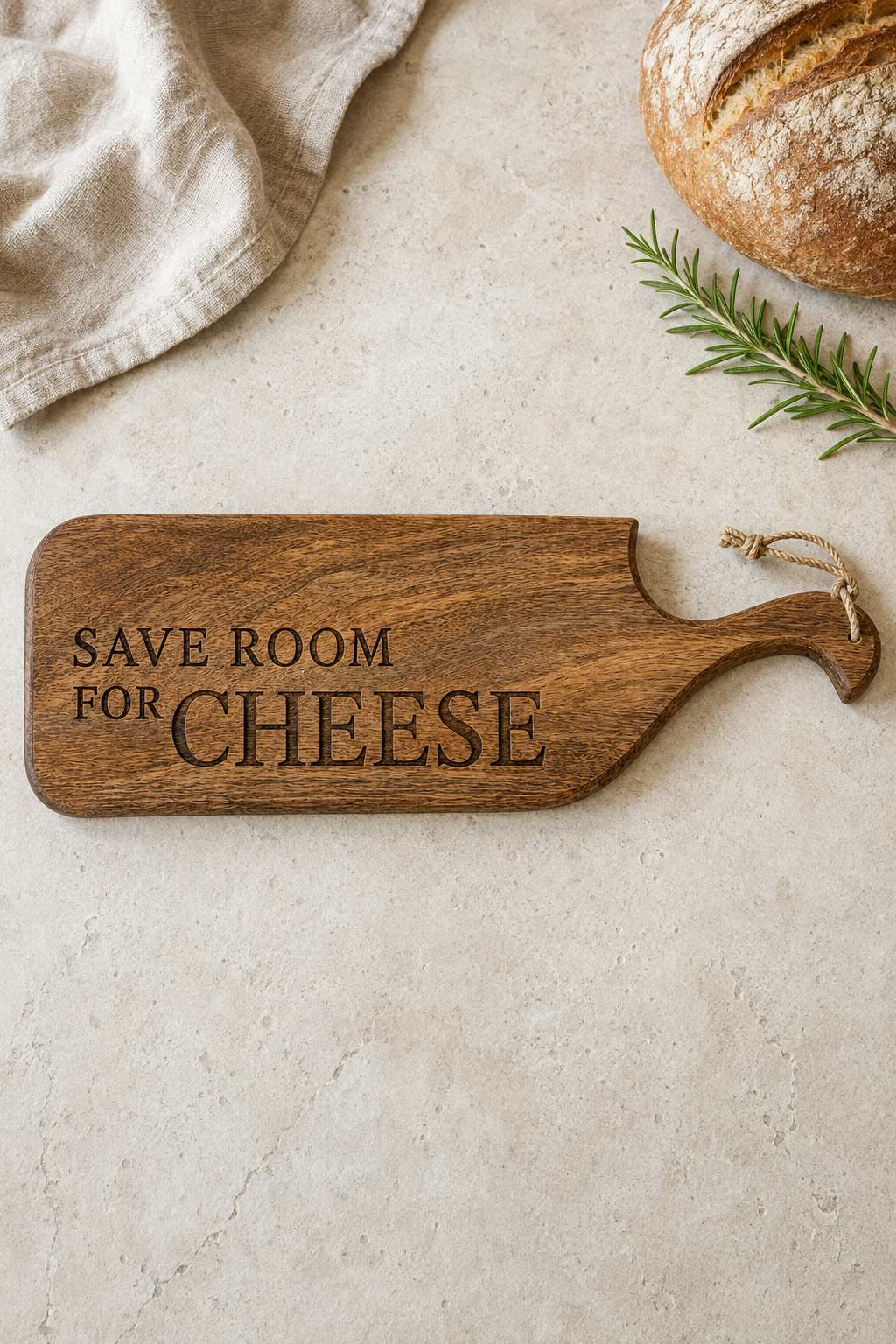 A wooden cheese board with the engraved phrase 'SAVE ROOM FOR CHEESE' and a hanging loop, styled on a light stone surface with a loaf of bread, a green sprig, and a folded cloth.