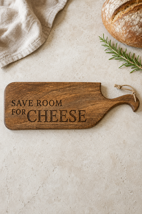 A wooden cheese board with the engraved phrase 'SAVE ROOM FOR CHEESE' and a hanging loop, styled on a light stone surface with a loaf of bread, a green sprig, and a folded cloth.