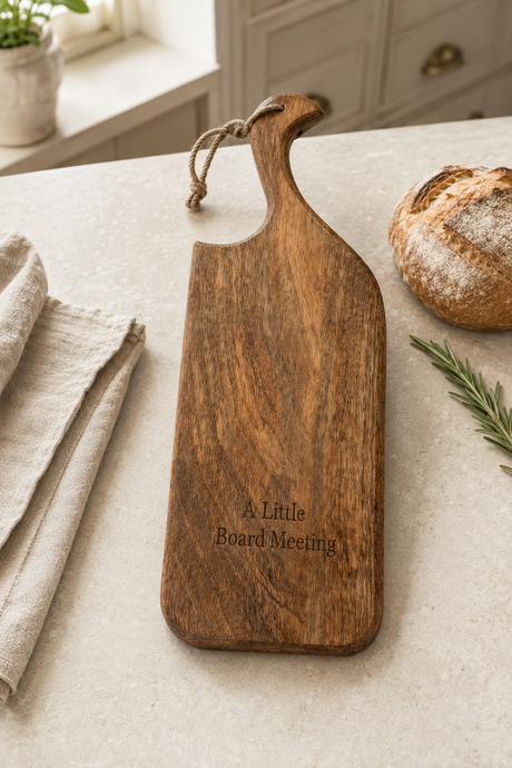 A wooden serving board with a rope handle and engraved text reading 'A Little Board Meeting' sits on a light stone countertop in a kitchen setting, with bread, a napkin, and rosemary nearby.