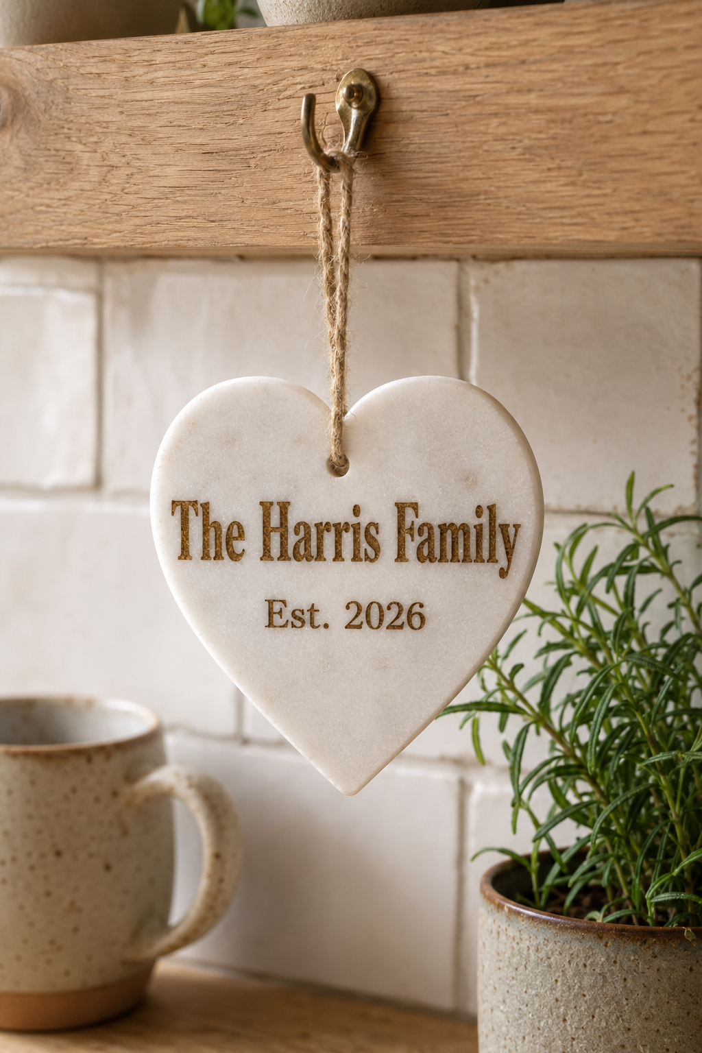 A personalised marble heart-shape hanging plaque engraved with 'The Harris Family Est. 2026' in brown lettering, suspended from a wooden shelf hook in a kitchen setting with a mug and potted plant nearby.