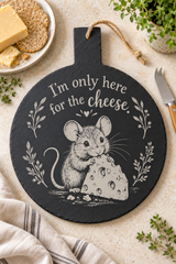 A round black slate cheese board with a hanging loop, featuring a white engraved illustration of a mouse holding cheese and the phrase 'I'm only here for the cheese.' The board is styled on a light stone surface with crackers, cheese, a knife, herbs, and a cloth napkin.
