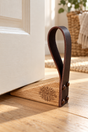 A wooden and leather door stopper resting on a light wood floor in front of a white door, with a woven basket and plant visible in the softly lit background.