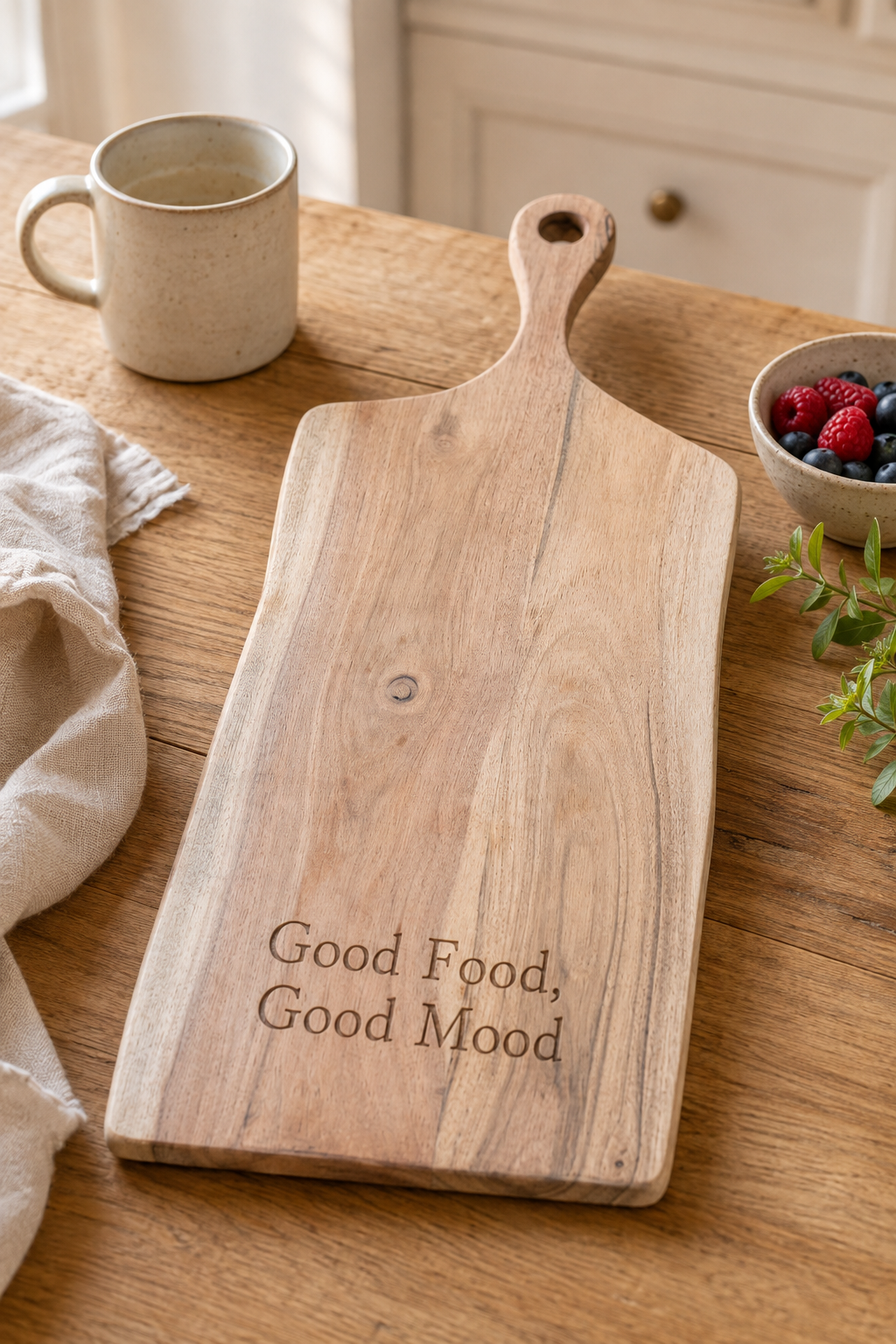 A rectangular wooden serving board with a handle, engraved with the phrase 'Good Food, Good Mood', displayed on a wooden table with a ceramic mug, a napkin, and a small bowl of berries.