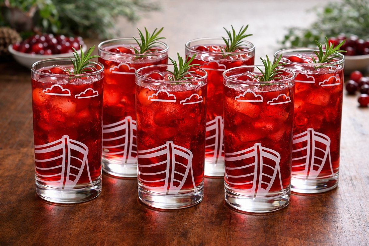 Set of six red cocktails with ice and rosemary in glasses with boat design etched on the glasses sat on a wooden surface.