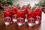 Set of six red cocktails with ice and rosemary in glasses with boat design etched on the glasses sat on a wooden surface.