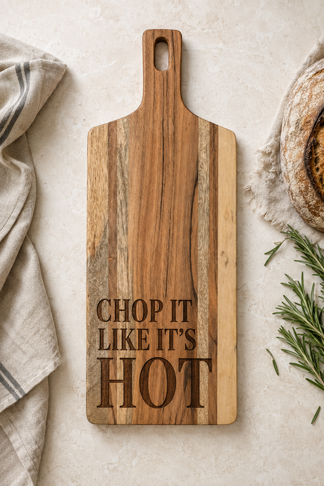 A rectangular wooden cutting board with a handle, featuring the engraved text 'CHOP IT LIKE IT'S HOT' in dark brown. The board is styled on a light beige textured surface with a striped kitchen towel, a loaf of bread, and rosemary.