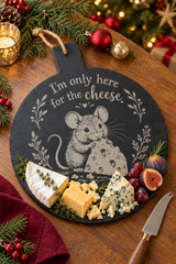A round black slate cheese board with a handle, engraved with a mouse holding cheese and the phrase 'I'm only here for the cheese.' The board is styled on a wooden table with cheeses, grapes, figs, a small knife, evergreen branches, and holiday ornaments in the background.