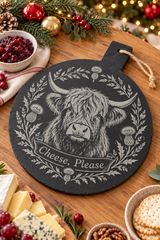 A round black slate cheese board with a white engraved Highland cow, thistle wreath, and the words 'Cheese, Please', styled on a wooden table with cheese, cranberries, nuts, and holiday decorations.