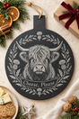 A round black slate cheese board with a white engraved illustration of a Highland cow surrounded by thistles and the words 'Cheese, Please'. The plaque is styled in a cozy holiday kitchen setting with cheese, crackers, dried orange slices, nuts, and a wrapped gift box.