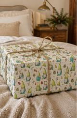 A gift box wrapped in cream wrapping paper with a repeating pattern of colorful gnomes and small trees, tied with a natural jute twine bow, resting on a cozy beige knit blanket in a bedroom setting.