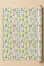 A roll of wrapping paper with a repeating pattern of cute green and blue gnomes on a cream background, displayed flat with the roll standing on the right side against a warm beige backdrop.